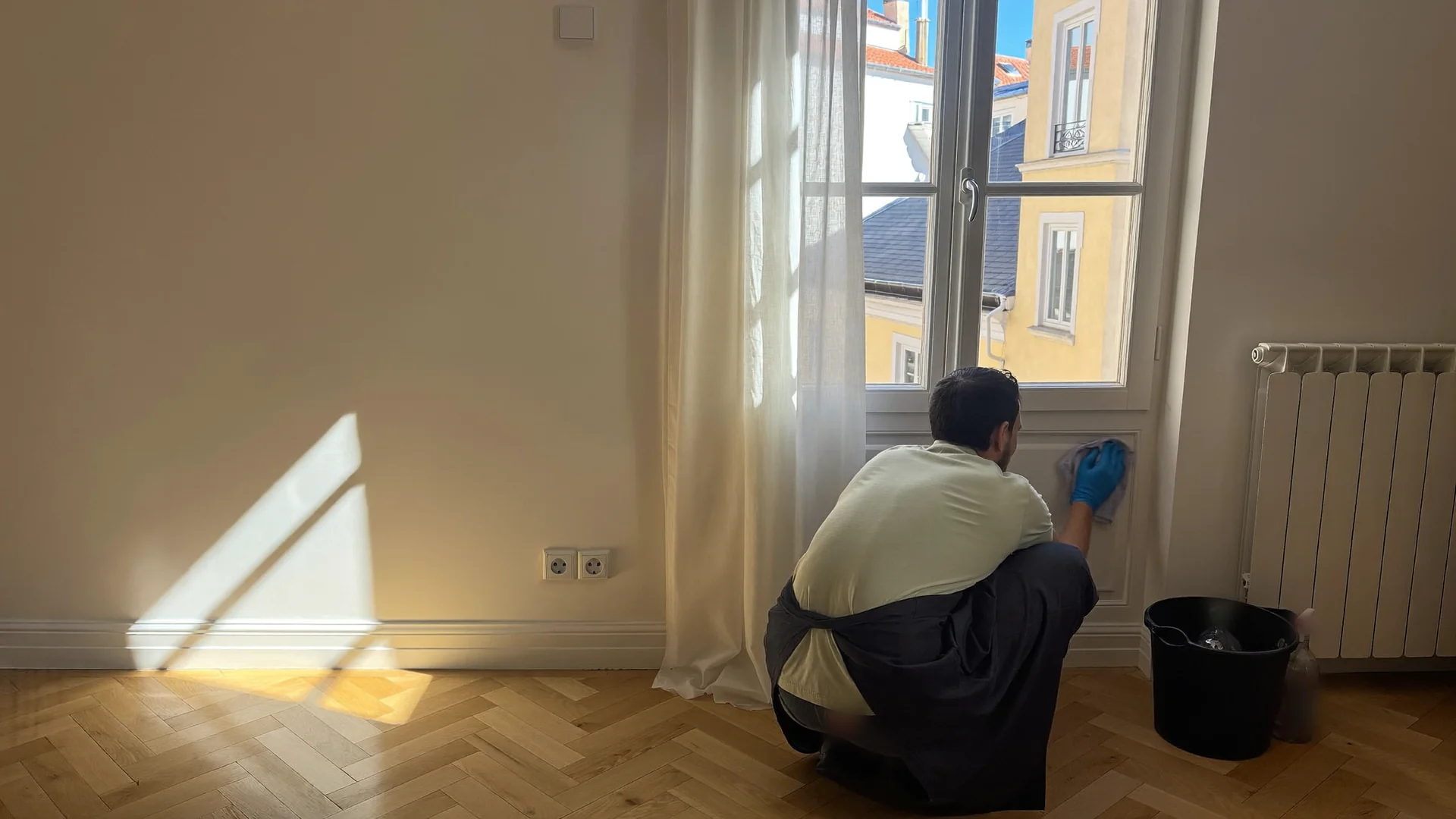 Cleaner in a work polo and gloves wiping a window frame in a sunlit Lisbon flat